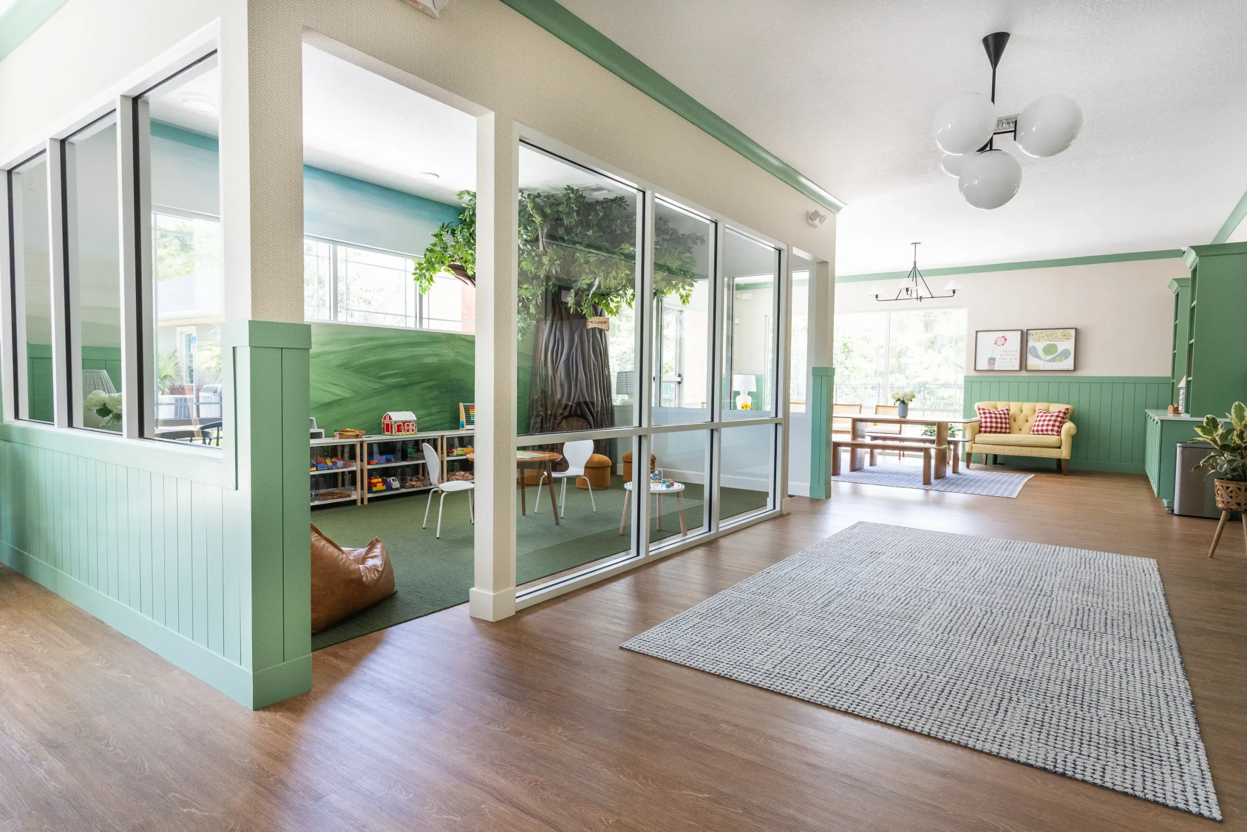 View into a glass-walled children's playroom with a tree mural and kid-sized furniture, beside a sunlit lobby seating area