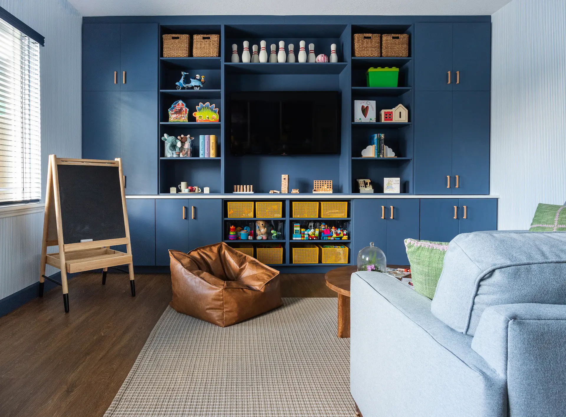 Children's playroom with navy blue built-in shelves holding toys and books, a chalkboard easel, leather bean bag, and gray sofa